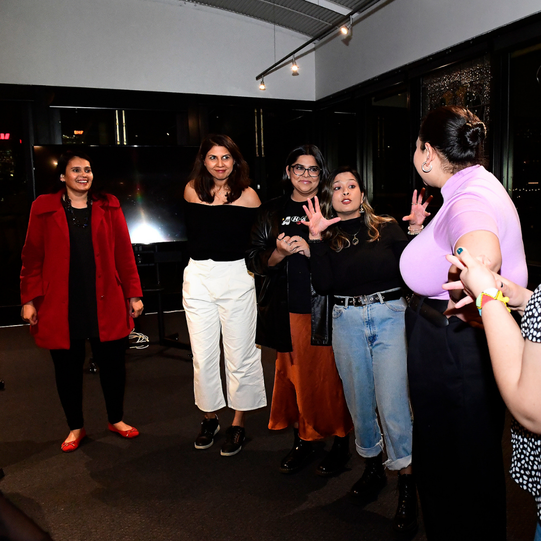 A group of women comedians stand in a circle during a workshop, laughing and interacting animatedly.